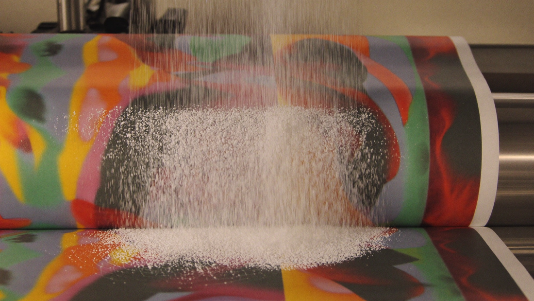 Automatic powder shaker applying adhesive powder onto a colorful printed DTF transfer film as it moves through the inline production line