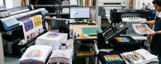 A print shop running sublimation printer and a DTF printer  with finished shirts on white and black fabric and an operator applying a DTF transfer at the heat press.