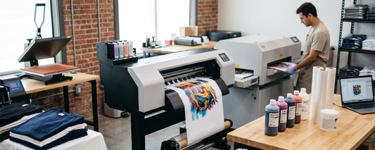 A t-shirt printing shop equipped with production machines, including a DTF printer and a DTG printer being operated by a technician in the background.